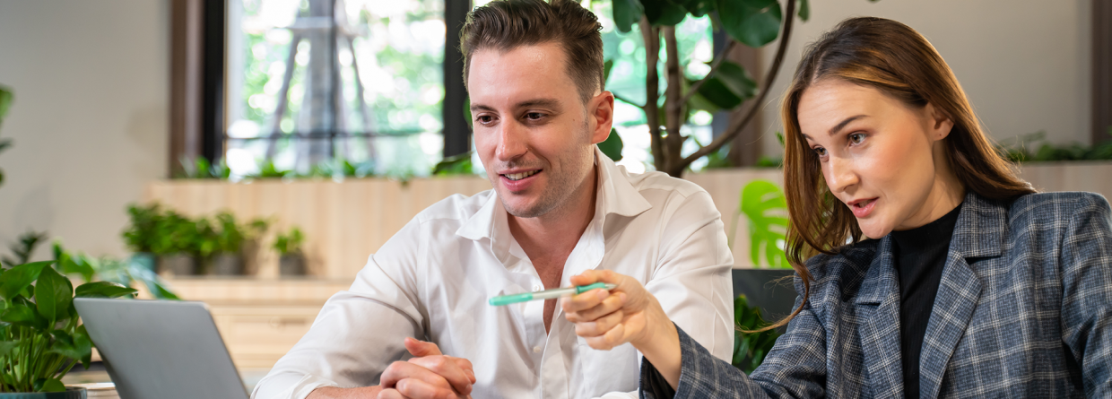 Bijscholing; een vrouw geeft begeleiding en uitleg aan een man op kantoor achter zijn bureau en laptop